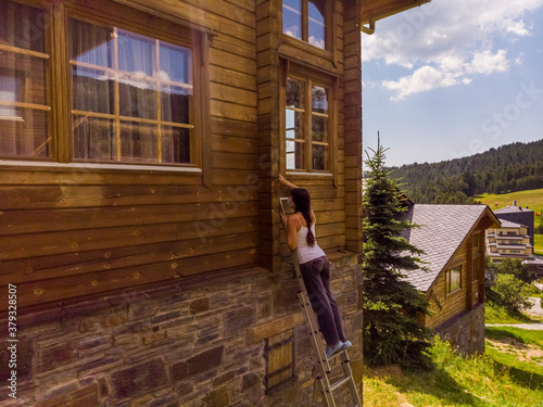 Woman on ladder with ponytail doing housework on holidays and varnishing and painting the wall of a chalet in the mountains