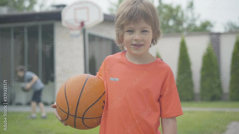 Little boy looking at camera and smiling as blurred kid playing basketball at the background. Portrait of cute Caucasian child posing outdoors with ball. Sportive childhood. Little boy looking at camera and smiling as blurred kid playing basketball at the background. Portrait of cute Caucasian child posing outdoors with ball. Sportive childhood.