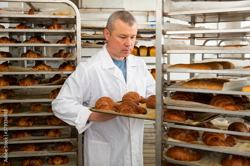 Portrait of positive baker working in bakehouse, putting tray with fresh bakery goods on rack trolley..