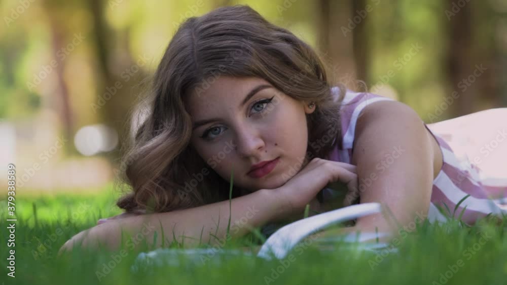 Young woman with curly fair hair rests lying on green grass near opened thick book and looks straight leaning on hand in park on summer day, slow motion closeup Young woman with curly fair hair rests lying on green grass near opened thick book and looks straight leaning on hand in park on summer day, slow motion closeup