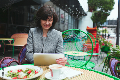 Mature woman sit alone in cafe or restaurant. Adult businesswoman hold tablet and work on it. Sit at table during lunch tinme eating salad. Working on project alone. photo