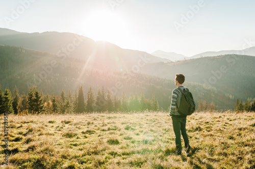 Young tourist man with a backpack on top of enjoying the sunset. Back view. Hipster walks in the autumn grass in the mountains on vacation. Holiday trip concept. World Tourism Day. © Serhii