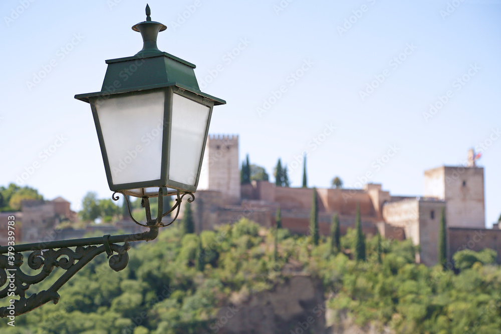 In the foreground a street lamp, in the background (out of focus) view of the Alhambra the ancient arabic fortress located in Granada, Spain