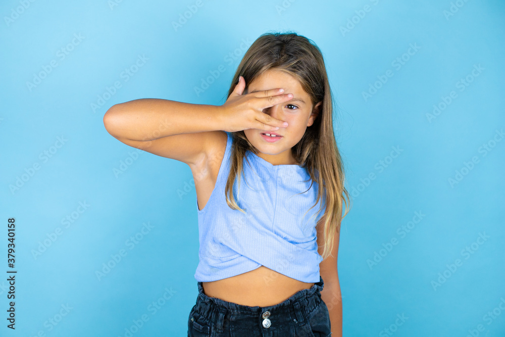 Young beautiful child girl over isolated blue background peeking in shock covering face and eyes with hand, looking through fingers with embarrassed expression Young beautiful child girl over isolated blue background peeking in shock covering face and eyes with hand, looking through fingers with embarrassed expression