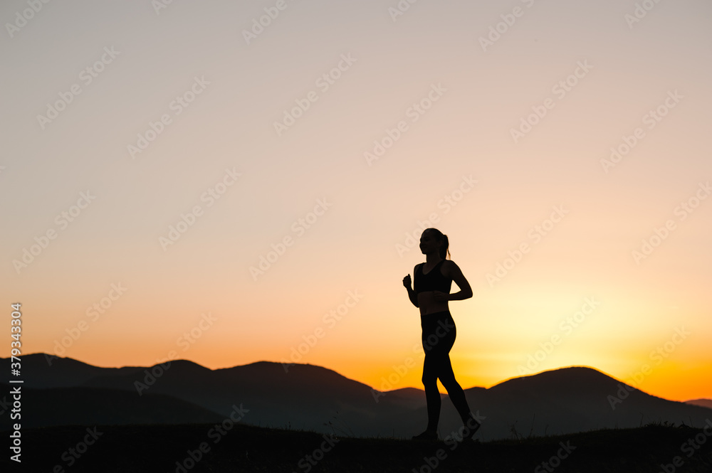 Athlete woman running on the road in the mountains. Silhouette against the sky at sunrise while running. Runner fitness girl in sport tight clothes. Bright sunset and blurry background. Horizontal.