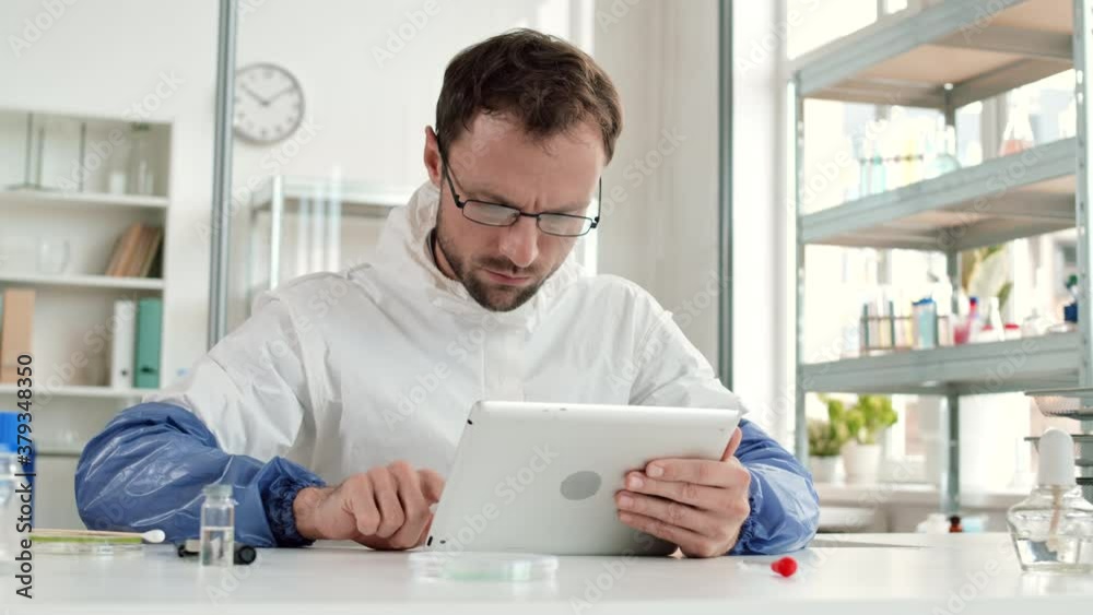 Medium shot of concentrated male researcher wearing coverall is sitting at desktop in modern laboratory and working on tablet Medium shot of concentrated male researcher wearing coverall is sitting at desktop in modern laboratory and working on tablet
