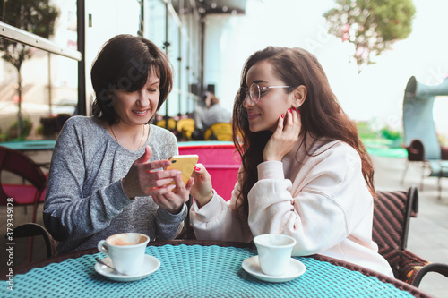 Mature mother and her young daughter sit together in cafe or restaurant. Girl shows her mother something on yellow smartphone. Watching together and having fun. Using modern technologies.