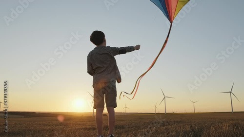 Happy boy launches flying bright kite into sky on mown wheat field, playing wind in field of an orange sunset on day lens flares wind turbines in summer slow motion. School break. Lifestyle. Childhood