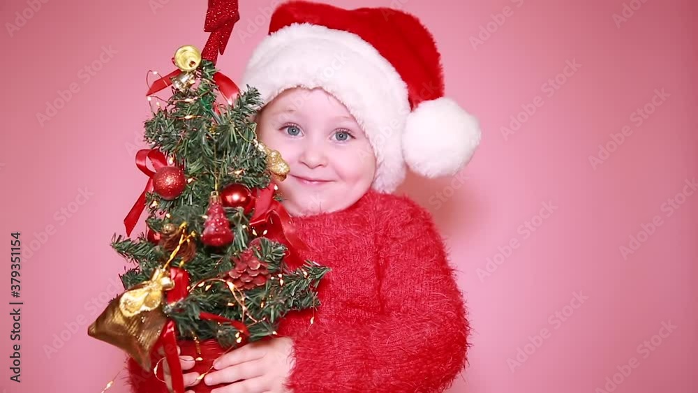 A little girl is sitting in a Santa Claus hat, holding a Christmas tree, smiling funny and looking at the camera. Isolate on a pink background