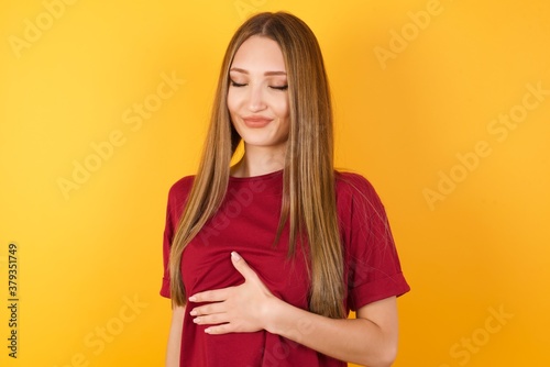 Beautiful Young beautiful caucasian girl wearing red t-shirt over isolated yellow background touches tummy, smiles gently, eating and satisfaction concept.