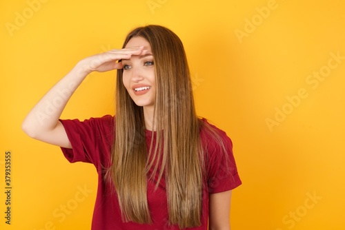 Beautiful Young beautiful caucasian girl wearing red t-shirt over isolated yellow background very happy and smiling looking far away with hand over head. Searching concept.