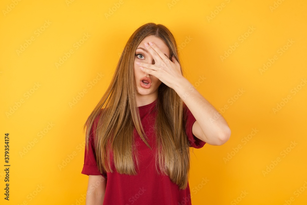 Beautiful Young beautiful caucasian girl wearing red t-shirt over isolated yellow background peeking in shock covering face and eyes with hand, looking through fingers with embarrassed expression. Beautiful Young beautiful caucasian girl wearing red t-shirt over isolated yellow background peeking in shock covering face and eyes with hand, looking through fingers with embarrassed expression.
