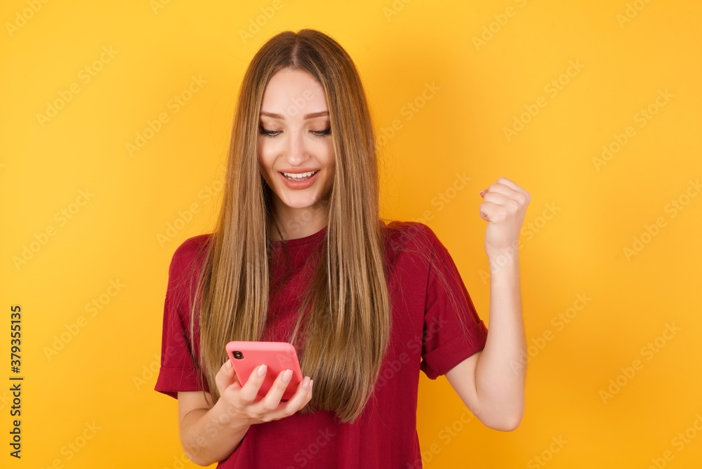 Beautiful Young beautiful caucasian girl wearing red t-shirt over isolated yellow background holding in hands cell reading browsing Beautiful Young beautiful caucasian girl wearing red t-shirt over isolated yellow background holding in hands cell reading browsing