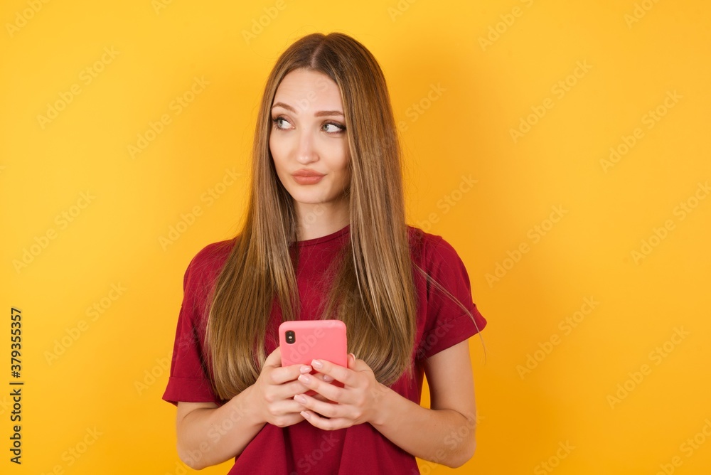 Funny Beautiful Young beautiful caucasian girl wearing red t-shirt over isolated yellow background holds telephone hands reads good youth news look empty space advert