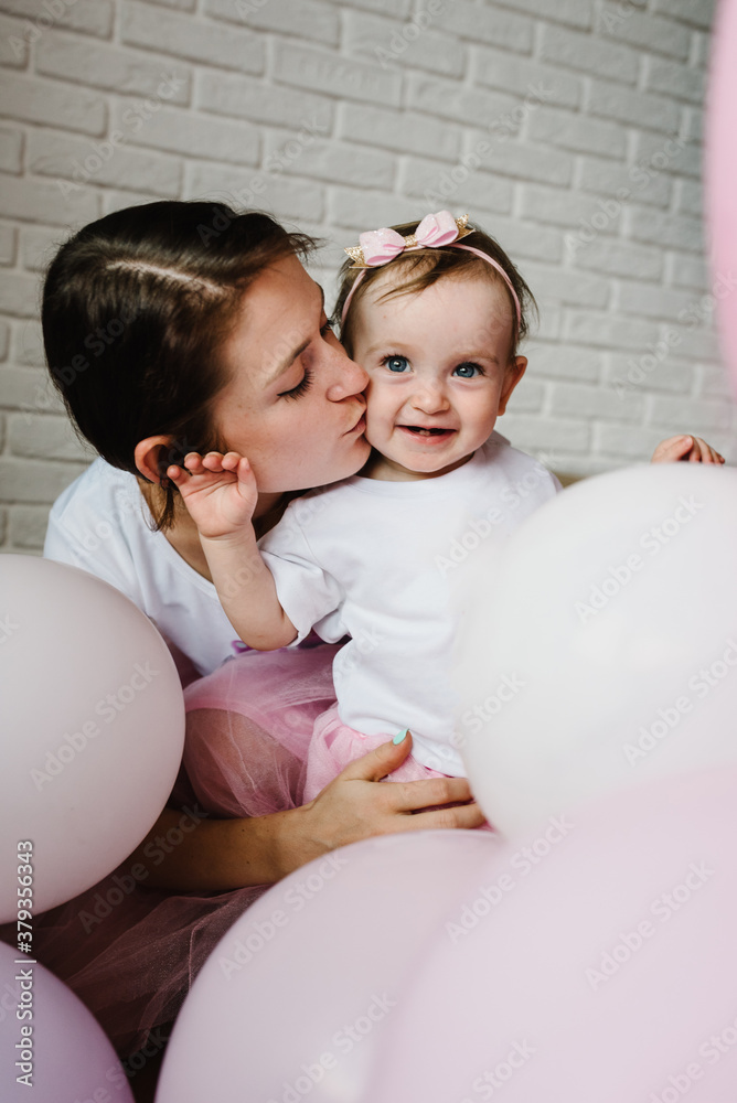 The mother holds little girl on background balloons indoors. Portrait of mom kissing daughter. The concept of family holiday. upper half. Close up. The mother holds little girl on background balloons indoors. Portrait of mom kissing daughter. The concept of family holiday. upper half. Close up.
