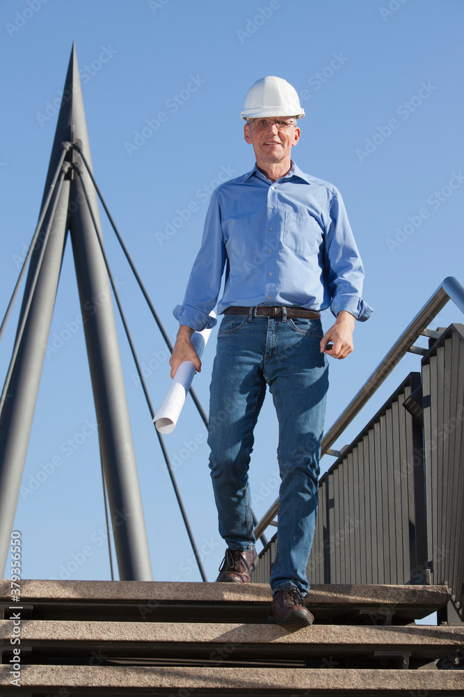 Smiling architect or engineer with hard hat and blueprint in his hand walking downstairs at a construction site Smiling architect or engineer with hard hat and blueprint in his hand walking downstairs at a construction site