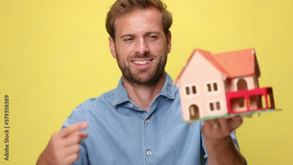 businessman holding a house in one hand and pointing at it with the other one, giving a thumbs up on yellow background businessman holding a house in one hand and pointing at it with the other one, giving a thumbs up on yellow background