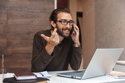 Smiling man with beard wearing glasses talking on cell phone while working on laptop