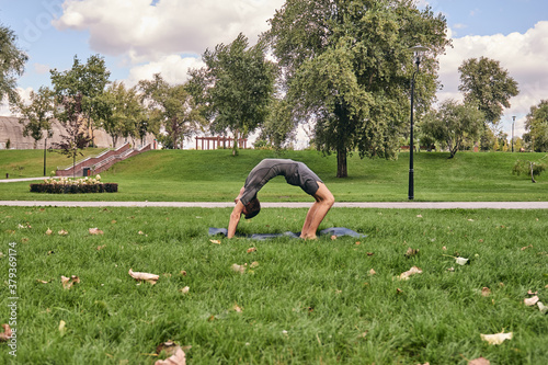 Young athletic man in sportswear doing yoga in the park. Practice asana outdoors. People exercising on green grass with yoga mat. Strong mature caucasian man in wheel pose