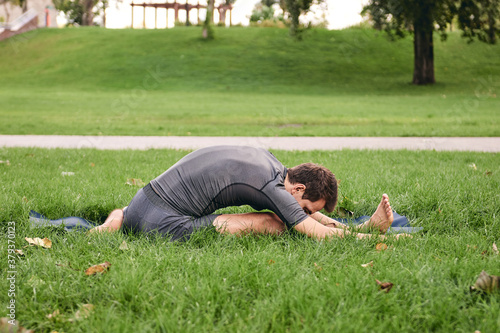 Young athletic man in sportswear doing yoga in the park. Practice Janu Sirshasana asana head to knee pose outdoors. People exercising stretching on green grass with yoga mat. © OneWellStudio