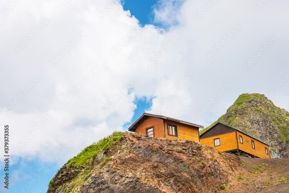 Two wooden houses on top of a mountain. Look up to the mountain range with clouds and sky.