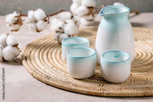 Tableau sur toile Sake ceramic set for traditional japanese alcohol drink rice wine sake, pitcher and three cups, standing on straw napkin with cotton flowers over beige texture background