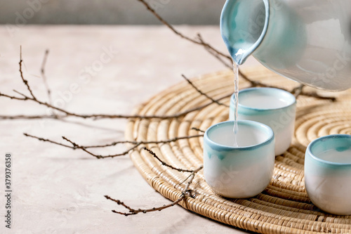 Sake ceramic set for traditional japanese alcohol drink rice wine sake pouring from pitcher in three cups, standing on straw napkin with dry branches over beige texture background.