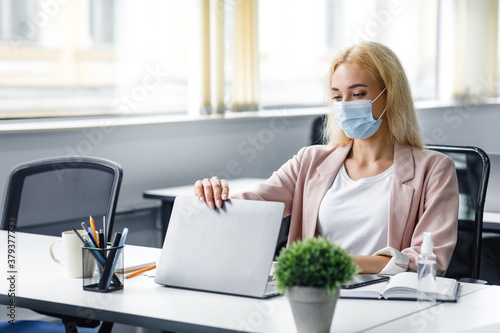 Modern workplace in coworking office with antiseptic. Beautiful businesswoman with medical mask working in office sitting at table with laptop