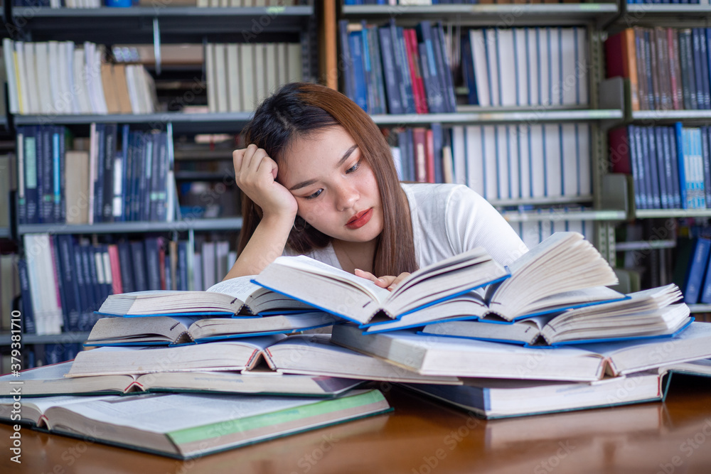 Asian female students are tired of reading in the library. A teenage female student sits on a table with a pile of books placed in front of them. Concept of reading books, boredom, test preparation Asian female students are tired of reading in the library. A teenage female student sits on a table with a pile of books placed in front of them. Concept of reading books, boredom, test preparation