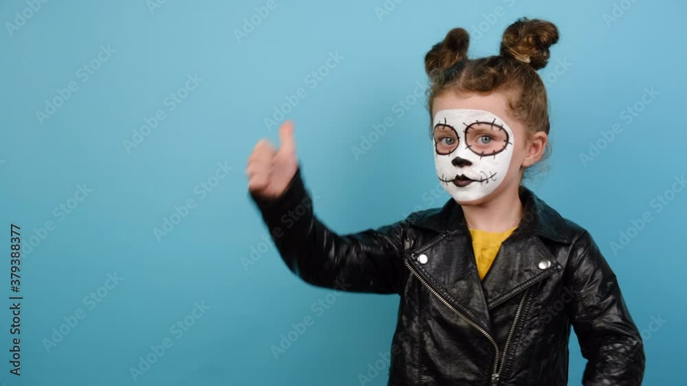Day of Dead and Halloween concept. Cute little girl with bright scary makeup, dressed in black leather jacket, points aside on blue copy space, showing okay hand sign, completely agree and support Day of Dead and Halloween concept. Cute little girl with bright scary makeup, dressed in black leather jacket, points aside on blue copy space, showing okay hand sign, completely agree and support