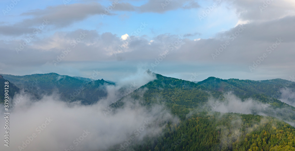 Fog in the mountains. Green and yellow forest on the mountain slopes. Wet fog in the valley. Sky with clouds. Takmak mountain in park Krasnoyarsk pillars. Natural blur.