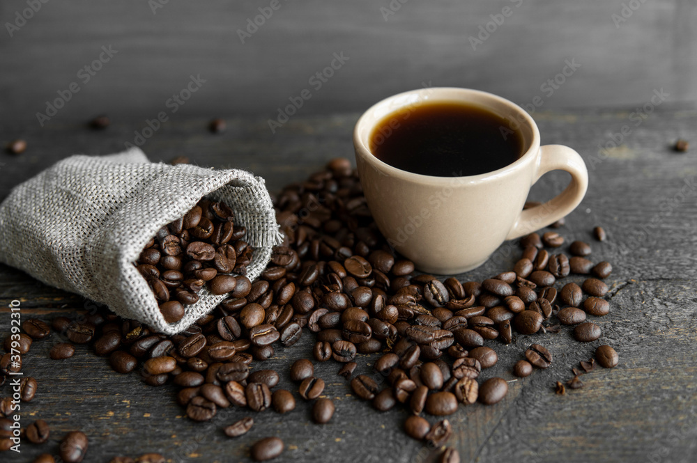 Coffee scattered fron a linen bag and a cup of black coffee Espresso or Americano standing on a wooden table. Fresh arabica coffee beans on linen textile. Aromatic freshly brewed coffee. Coffee scattered fron a linen bag and a cup of black coffee Espresso or Americano standing on a wooden table. Fresh arabica coffee beans on linen textile. Aromatic freshly brewed coffee.