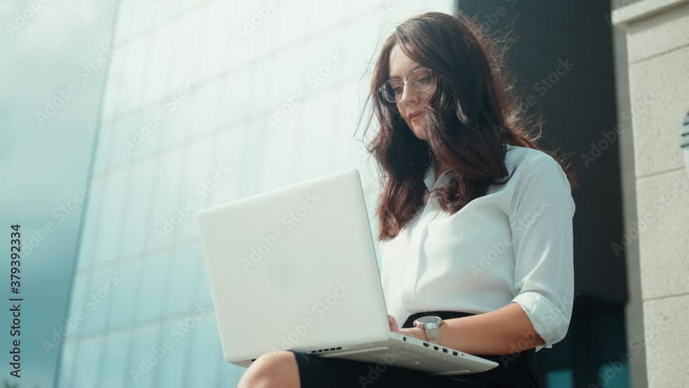 Remote work. A business woman works on a laptop, sitting comfortably on a bench by the business center Remote work. A business woman works on a laptop, sitting comfortably on a bench by the business center
