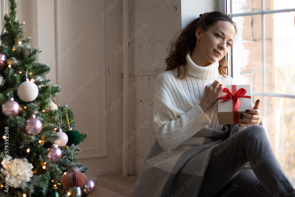 Beautiful young woman holding a christmas gift box and looking at the window thoughtfully next to the New Year tree. Cozy winter and autumn time. Beautiful young woman holding a christmas gift box and looking at the window thoughtfully next to the New Year tree. Cozy winter and autumn time.