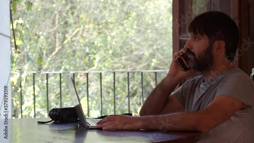 
Bearded man talking on the smartphone while working with a laptop in a house in the middle of nature.Digital nomad, content craver photo