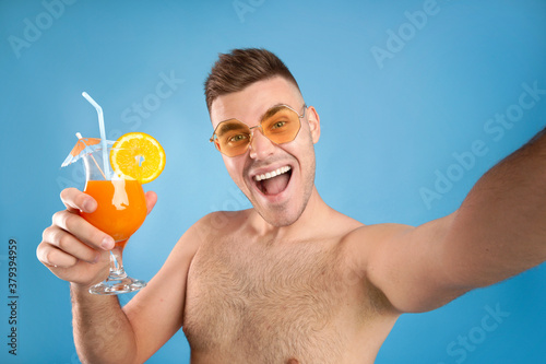Excited young man in sunglasses holding refreshing summer cocktail and taking selfie on blue studio background