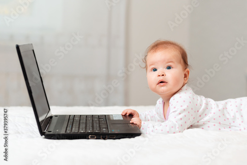 Cute baby is lying on the bed and touching the laptop. Side view. Concept of online communications