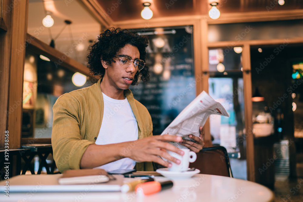Pensive male freelancer reading newspaper in cafe and drinking coffee