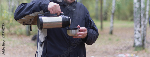 A European man in a black jacket pours hot tea from a thermos in a hike. A man wants to drink tea in the forest in autumn.