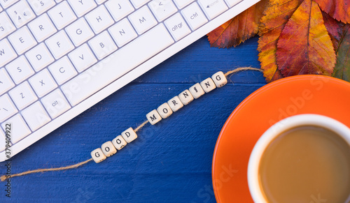 Desktop with White Computer English Keyboard, Orange Cup of Coffee Latte, Autumn Bright Leaves and Creative Text “Good Morning” – Top View. Autumn Mood on Workplace. 