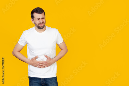 A young man with a beard in a white T-shirt with a hand on his stomach because of indigestion, feeling sick. Pain concept. Stands on isolated yellow background © Yauhen