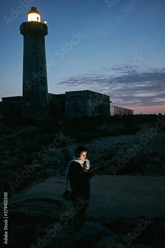 jeune femme regardant son smartphone dans la nuit en bord de mer à côté d'un phare en Sicile photo