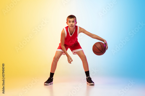 Full length portrait of young basketball player in uniform on gradient studio background. Teenager confident practicing with ball. Concept of sport, movement, healthy lifestyle, ad, action, motion. © master1305