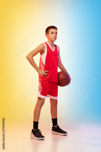 Full length portrait of young basketball player in uniform on gradient studio background. Teenager confident posing with ball. Concept of sport, movement, healthy lifestyle, ad, action, motion.