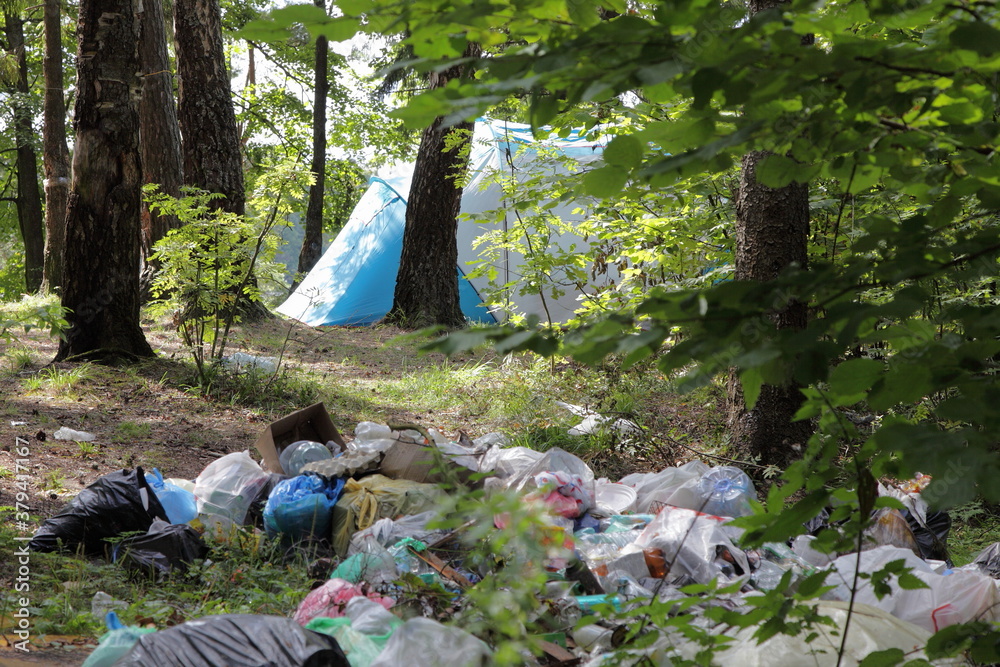 Big garbage pile in the forest next to a camp tent among the green trees in Park on a Sunny summer day, plastic littering the wildlife with vacationers people