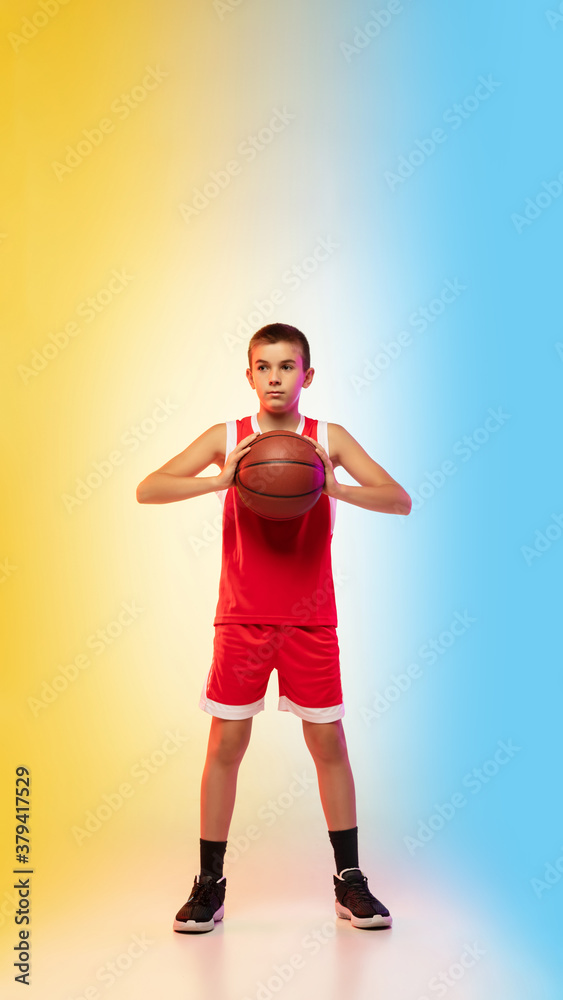 Full length portrait of young basketball player in uniform on gradient studio background. Teenager confident posing with ball. Concept of sport, movement, healthy lifestyle, ad, action, motion.