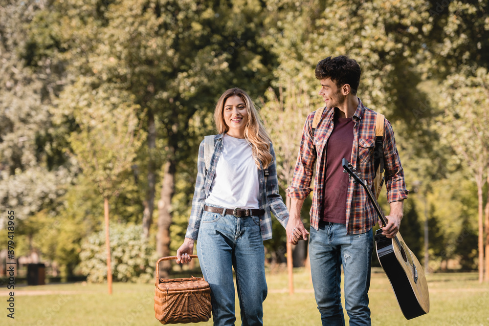 woman with wicker basket and man with acoustic guitar holding hands in park woman with wicker basket and man with acoustic guitar holding hands in park