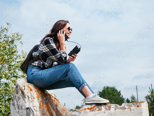 A happy girl in the mountains is laughing while talking on a phone charging from power bank. A girl in casual clothes on vacation shares her emotions. Concept for modern gadgets and portable chargers photo