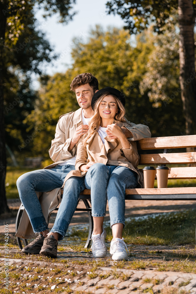 blonde woman in hat and man in trench coat sitting on wooden bench near paper cups