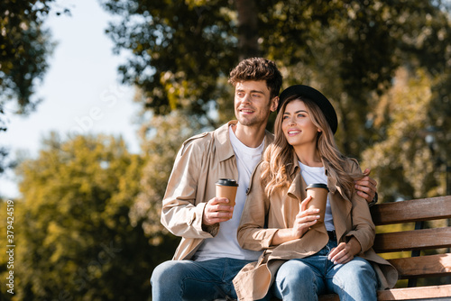 blonde woman in hat and man in trench coat holding disposable cups while sitting on bench